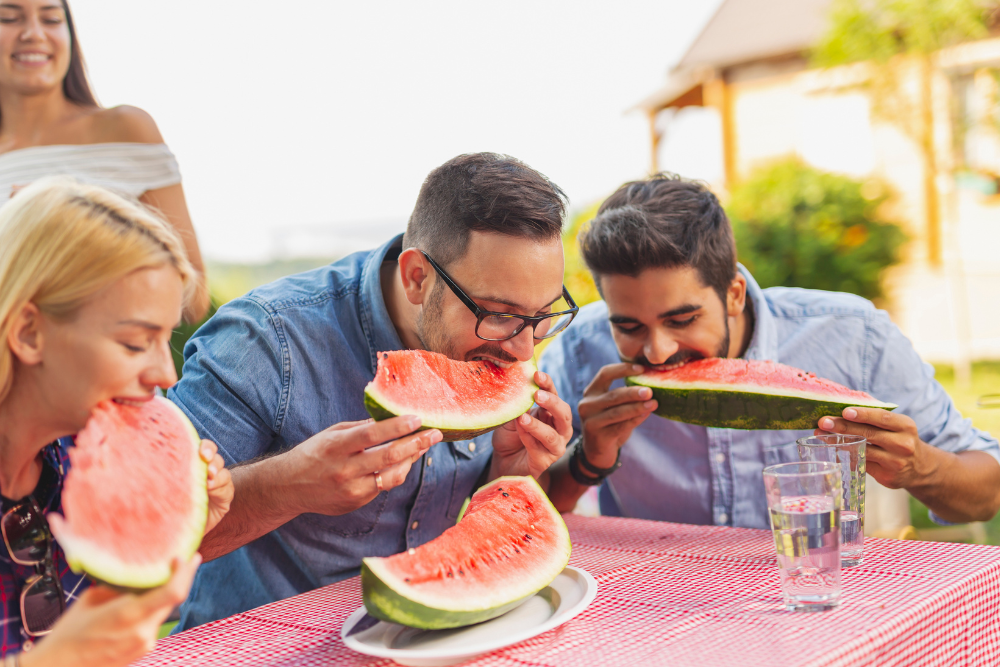 Two men eating watermelon slices outdoors at a table.